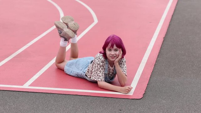 Young girl with bright pink hair and face glitter lies on stomach on pink outdoor sports court, propping head on hand and smiling toward camera, wearing denim overalls, leopard print shirt, white
