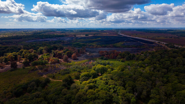 Aerial view of the New Forest National Park showing a wildfire burn scar near the A31 road, with green woodland under a cloudy sky in New Forest District, England, United Kingdom.
