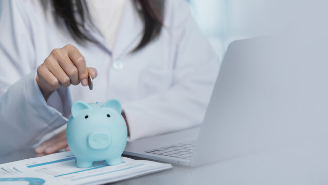 Doctor putting coin into blue piggy bank on office desk with laptop for medical investment, healthcare fund savings and insurance planning concept. Medical technology. hospital management.