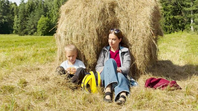 A mother and son sit and eat a snack by a large haystack in a sunny field. They feel happy, peaceful, and relaxed during this joyful outdoor break.