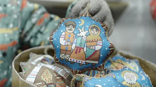Gloved hand lifts and presents padded star-shaped textile ornament decorated with folk-style embroidered scene of two figures and goat against bright blue background