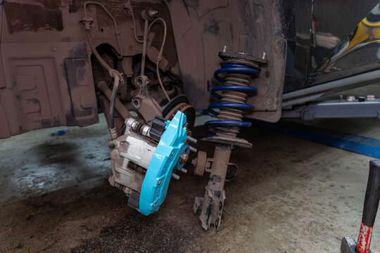 Brake repair with exposed caliper and rotor hub inside dirty wheel well. Blue part, spring strut and wheel studs stand out during maintenance on vehicle suspension with coil spring and wheel hub.