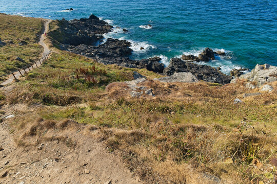 A scenic photo of a coastal path winding along a grassy cliff overlooking a rocky coastline and the turquoise sea - St Ives, Cornwall, UK
