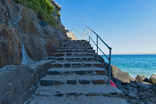 Stone steps climb a rocky cliff beside a bright blue ocean under a clear sky. A metal handrail assists the ascent - St Ives, Cornwall, UK