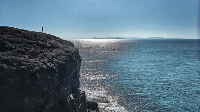 Aerial view of a person standing on a rugged volcanic cliff edge overlooking the sparkling blue Atlantic Ocean under a clear sky in Lanzarote, Canarias, Spain.