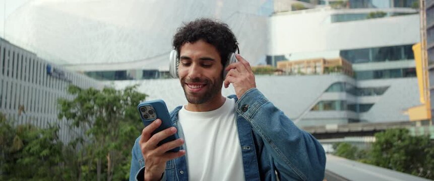 Portrait of happy Hispanic man listens to music using earphones and smartphone in city anamorphic lens. Latin guy sings enjoying favorite song in megapolis. Urban lifestyle