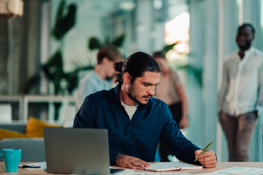 Focused businessman writing notes in a modern open plan office, working on laptop while colleagues move in background