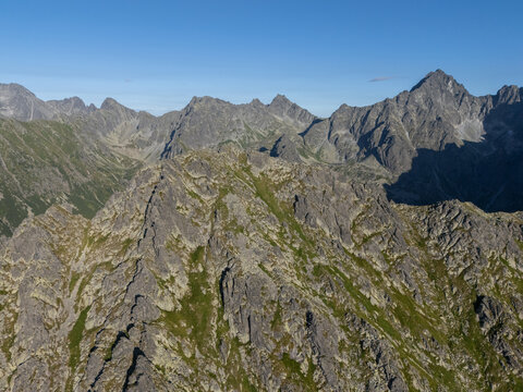 Aerial view of Mlynar peak rising above the rugged terrain of Bielovodska dolina with rocky slopes and green vegetation under a clear blue sky in Tatranska Javorina, Presov Region, Slovakia.