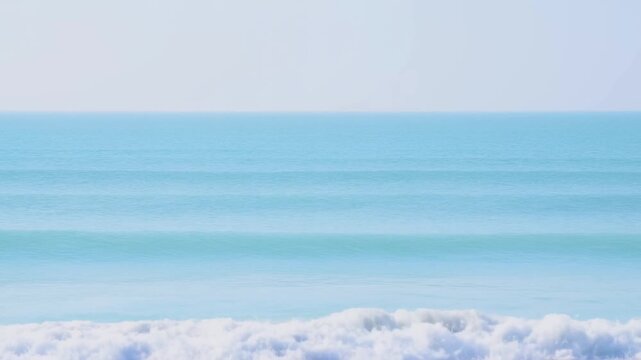 Slow motion static tracking pan of ocean waves rolling and breaking on sandy shoreline with horizon in soft morning light