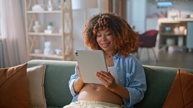 Smiling pregnant woman using tablet home sofa. Happy african american mother.