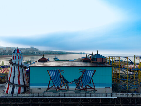 Aerial view of Brighton Palace Pier featuring a mural of two deckchairs on a building, with amusement rides, the sea, and a clear sky in Brighton and Hove, England, United Kingdom.