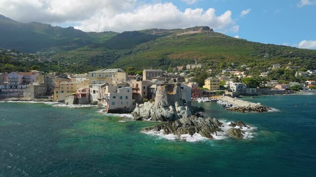 Erbalunga Cap Corse Corsica 4K drone video. Fly in from sea to ruined Genoese tower on rocky coast with crashing waves. Smooth turn reveal of old village houses overhanging water b roll