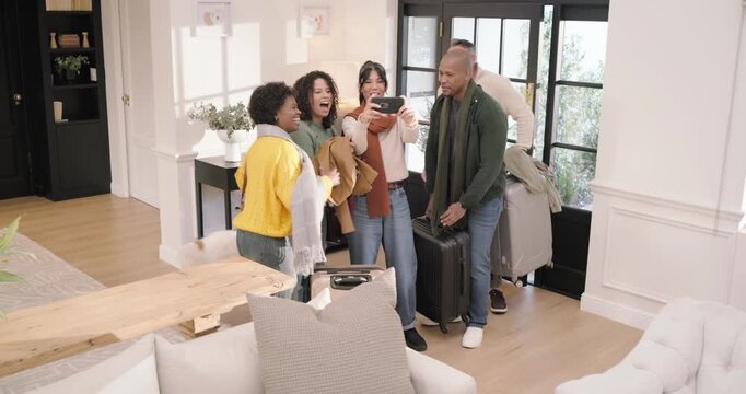 Diverse friends arriving through front door at home, dropping suitcases and taking selfies on phone
