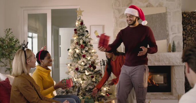Diverse friends sitting by Christmas tree exchanging gifts while Santa hat man handing parcels