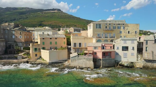 Erbalunga Cap Corse Corsica 4K drone view. Side tracking flyby of sunlit village houses overhanging emerald sea with mountains and turn reveal of ruined Genoese tower cape. Authentic Corsican b roll