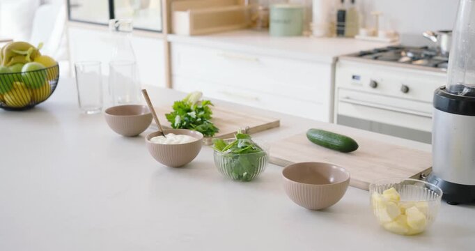 Camera is slowly panning left from right to show blender, cucumber, herbs, bowls, butter on counter
