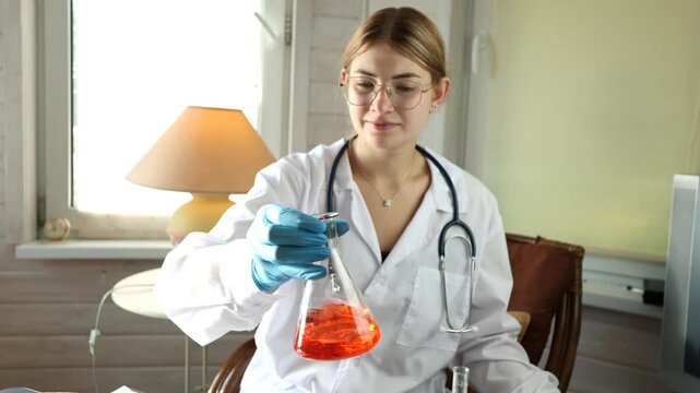 Female doctor in white coat gently swirls glass flask filled with orange red solution, then lowers it while keeping attention on liquid. Sunlit interior and table lamp create warm lab atmosphere.