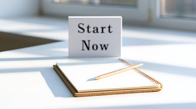 A motivational sign with text start now placed on a white desk next to an open notebook and pencil representing new goals and productivity