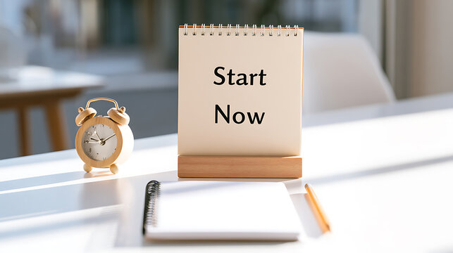 A motivational notebook display with start now text on a white office desk next to a small vintage alarm clock in a bright workspace