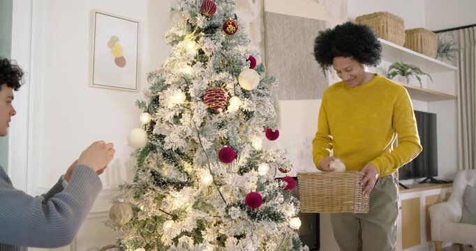 African American men decorating home for holidays, passing ornaments from basket on flocked tree