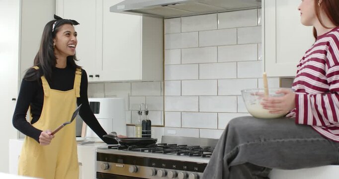 Diverse female friends cooking in kitchen after batter set holding bowl and tending pan with turner