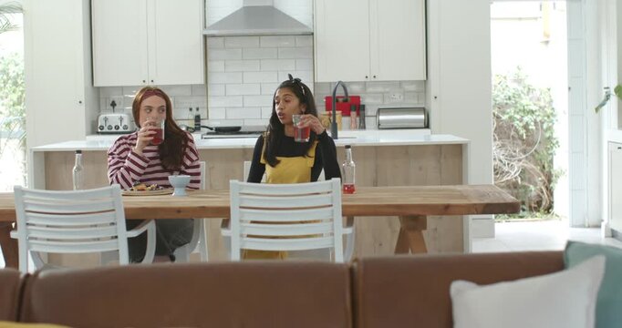 Diverse women sitting at table, right woman raising glass to start chat, left woman listening