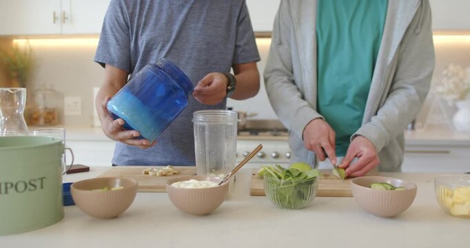 African American dad and teenage son pouring powder into blender at counter, chopping for smoothie