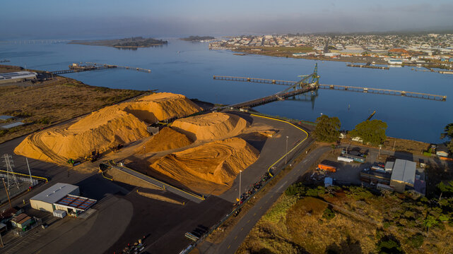 Aerial view of large wood chip piles at an industrial port facility with a pier extending into the bay in Samoa, California, United States.