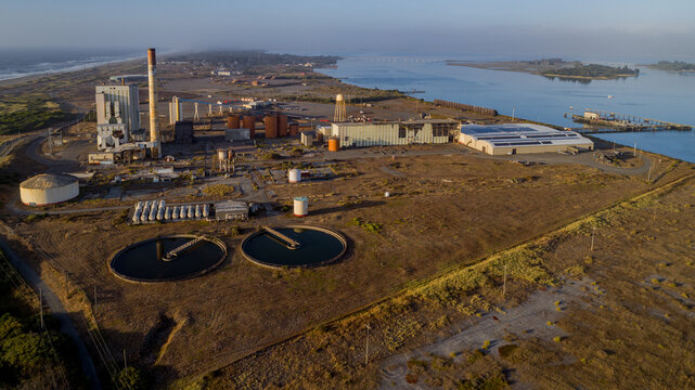 Aerial view of an industrial pulp mill facility featuring large storage tanks, a tall chimney, and circular sedimentation ponds near the coastline in Samoa, California, United States.