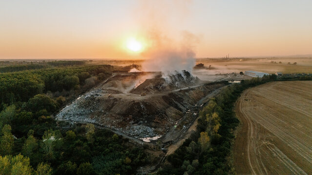 Aerial view of a landfill site with smoke rising from waste piles, surrounded by agricultural fields and forests during sunset in Sremska Mitrovica, Vojvodina, Serbia.