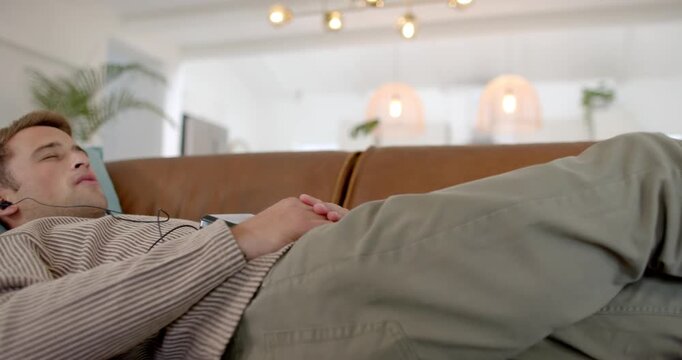 Camera pulling back man reclining on sofa adjusting hands, listening with wired earphones, relaxing