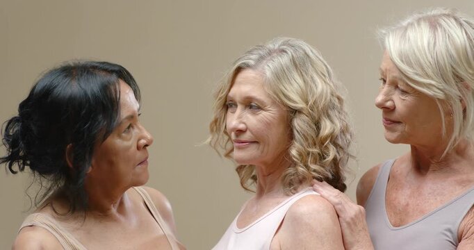 Mature women peers facing gaze with hand on shoulder, softening, turning forward on beige backdrop