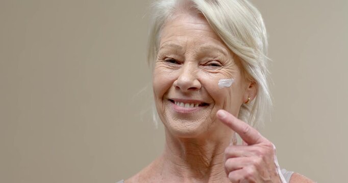 Senior woman touching cream with index finger on cheek, reacting to dab for skincare in studio