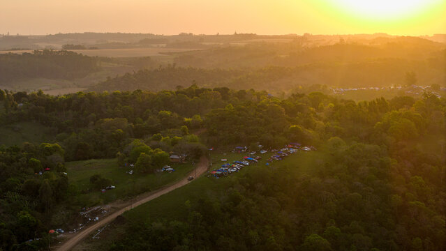 Aerial view of the Rally Paraguay 2026 spectator area with cars and tents along a dirt road amidst green hills during a golden sunset in Nueva Alborada, Itapua Department, Paraguay.