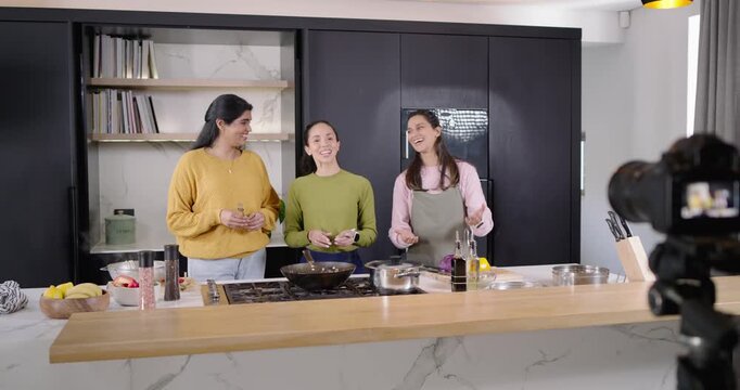Diverse women filming cooking in kitchen camera rolling reaching pan hugging waving one in apron