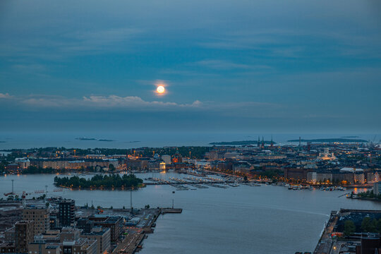 Aerial view of the Helsinki harbor at dusk with a full moon rising over the city skyline, illuminated buildings, and boats docked in the water in Helsinki, Uusimaa, Finland.
