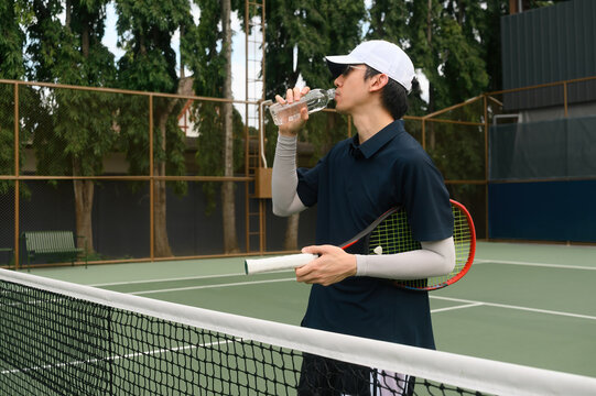 Young Asian Male Tennis Player Drinking Water on Outdoor Tennis Court During Break