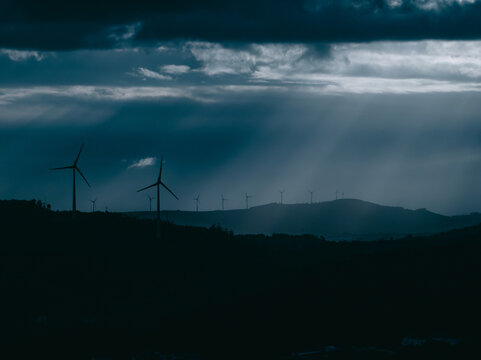 Aerial view of wind turbines on a hill under a dramatic dark sky with sun rays breaking through the clouds in Santo Isidoro, Lisboa, Portugal.