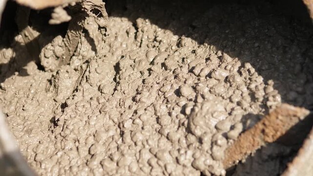 Wet concrete or mortar sits inside cylindrical container, showing coarse lumpy texture, glossy moisture, bright central highlights, deep rim shadow, and rugged construction detail in tight close view