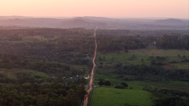 Aerial view of a straight dirt road cutting through rural fields, dense forests, and rolling hills under the warm golden hour light in Nueva Alborada, Itapua Department, Paraguay.