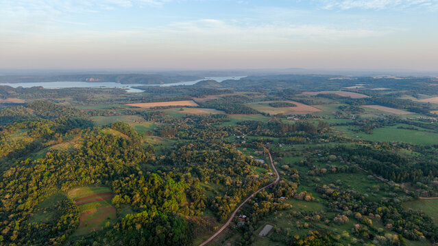 Aerial view of rolling hills, dense forests, and agricultural fields surrounding a winding dirt road in the countryside of Nueva Alborada, Itapua Department, Paraguay.
