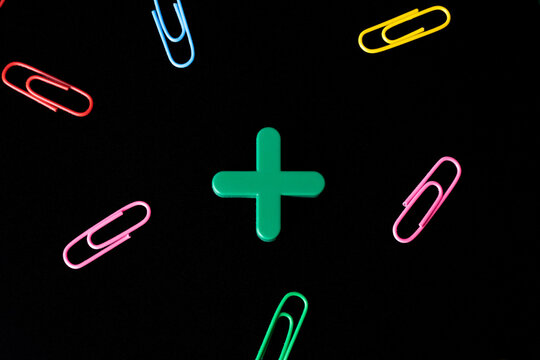 Green wooden plus symbol surrounded by scattered colorful paper clips on black background