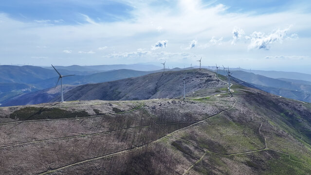 Aerial view of a wind farm with multiple wind turbines standing along a mountainous ridge, featuring dirt access paths and rolling hills under a bright blue sky in Lisbon, Lisbon, Portugal.