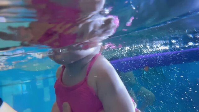 Toddler, baby girl in a pink swimsuit dives underwater, smiling with her eyes open. Underwater view of infant swimming, early childhood development, water confidence, swimming lessons and active