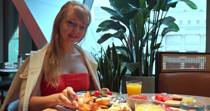 Caucasian woman in red dress eating breakfast at restaurant table. Pastries, fruit, and orange juice served in modern cafe interior with green plants. Morning dining lifestyle.