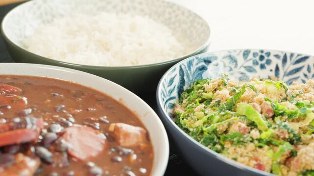Homemade Brazilian feijoada made with black beans and pieces of pork, served in bowls with white rice and toasted cassava flour. Traditional Brazilian lunch prepared at home.