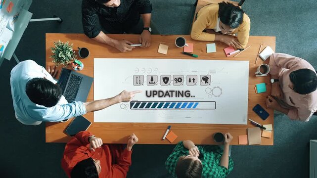 A diverse team collaborates on a project update in an office setting, discussing ideas and strategies around a long table filled with laptops and notes. Raster