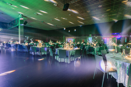 Elegant banquet hall setup with round tables adorned with green tablecloths and floral centerpieces, illuminated by colorful lights and a spacious dance floor in the background
