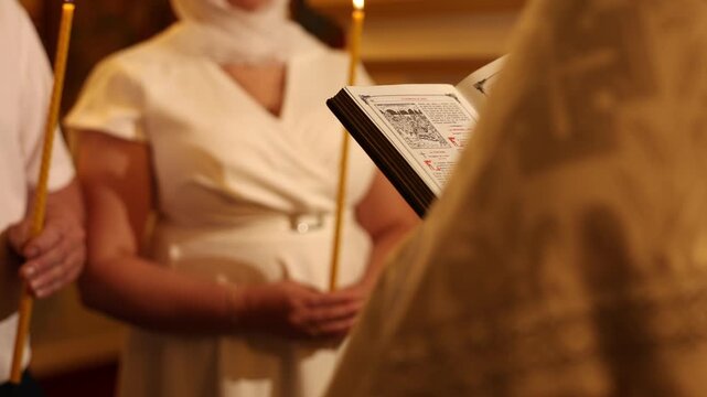 Close over-shoulder of reader presenting open book toward candlelit congregation during formal ceremony, warm amber lighting and shallow depth of field, with book, page, reading, reader detail