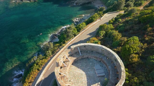 Tour de l Osse Cap Corse Corsica 4K drone video. Top down fly to winding road at sunset. Tracking flight along coastal highway over small beach and Genoese watchtower in golden hour light